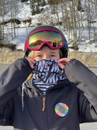 a girl in snow gear pulling a voltage neck sock up over her face