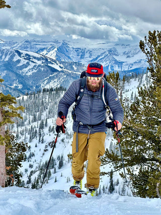 A man hiking up a snowy mountain with ski poles in an iron man hat