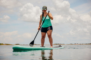 A woman paddle boarding on a lake wearing a Voltage Trucker hat